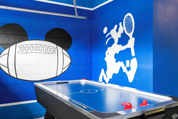 Een kamer met een tafelvoetbaltafel, een basketbalring en een muurschildering met een tennisracket en een voetbal.