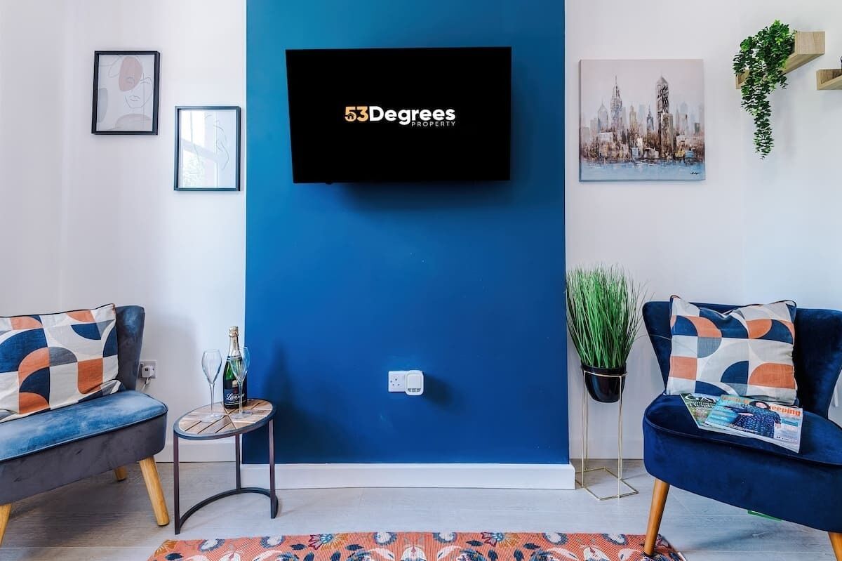 A modern living room with a blue accent wall, two sofas with patterned cushions, a flat-screen TV, and a small side table with a bottle and glasses.