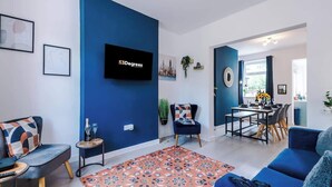 Un saló modern amb una paret d'accent blava, un televisor de pantalla plana, una zona de menjador amb taula i cadires, i una catifa amb dibuixos.