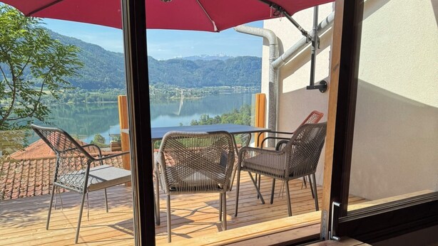 Eine Holzterrasse mit Gartenmöbeln, einem roten Sonnenschirm und Blick auf einen See und Berge.
