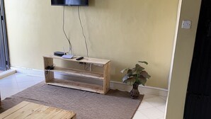 Un salon avec un téléviseur fixé au mur, un banc en bois, une petite table, une plante en pot et un sol carrelé.
