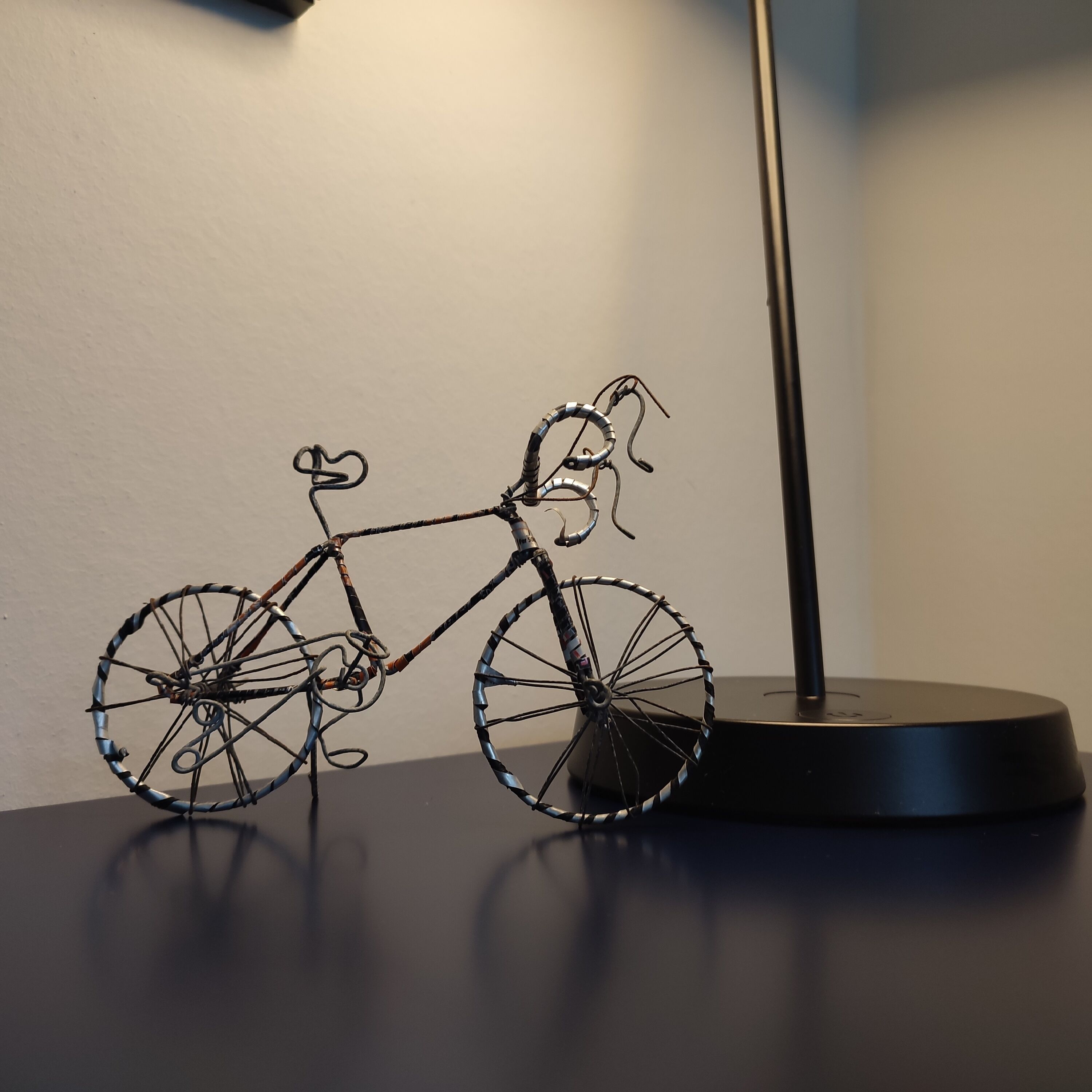 Una scultura in filo a forma di bicicletta su una superficie scura, accanto a una lampada.