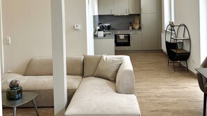 A modern living room with a beige sectional sofa, a small coffee table, and a black metal chair. A kitchen with white cabinets and a stove is visible in the background. - Pure Highlight Living 9 - Holiday apartment / temporary living (Steinfurt)