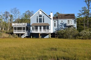 A large two-story house with a balcony and a covered porch, surrounded by a grassy field and trees. - Prospect Point- 4BR Waterfront | Screened Porch | Private Pier (Chincoteague)