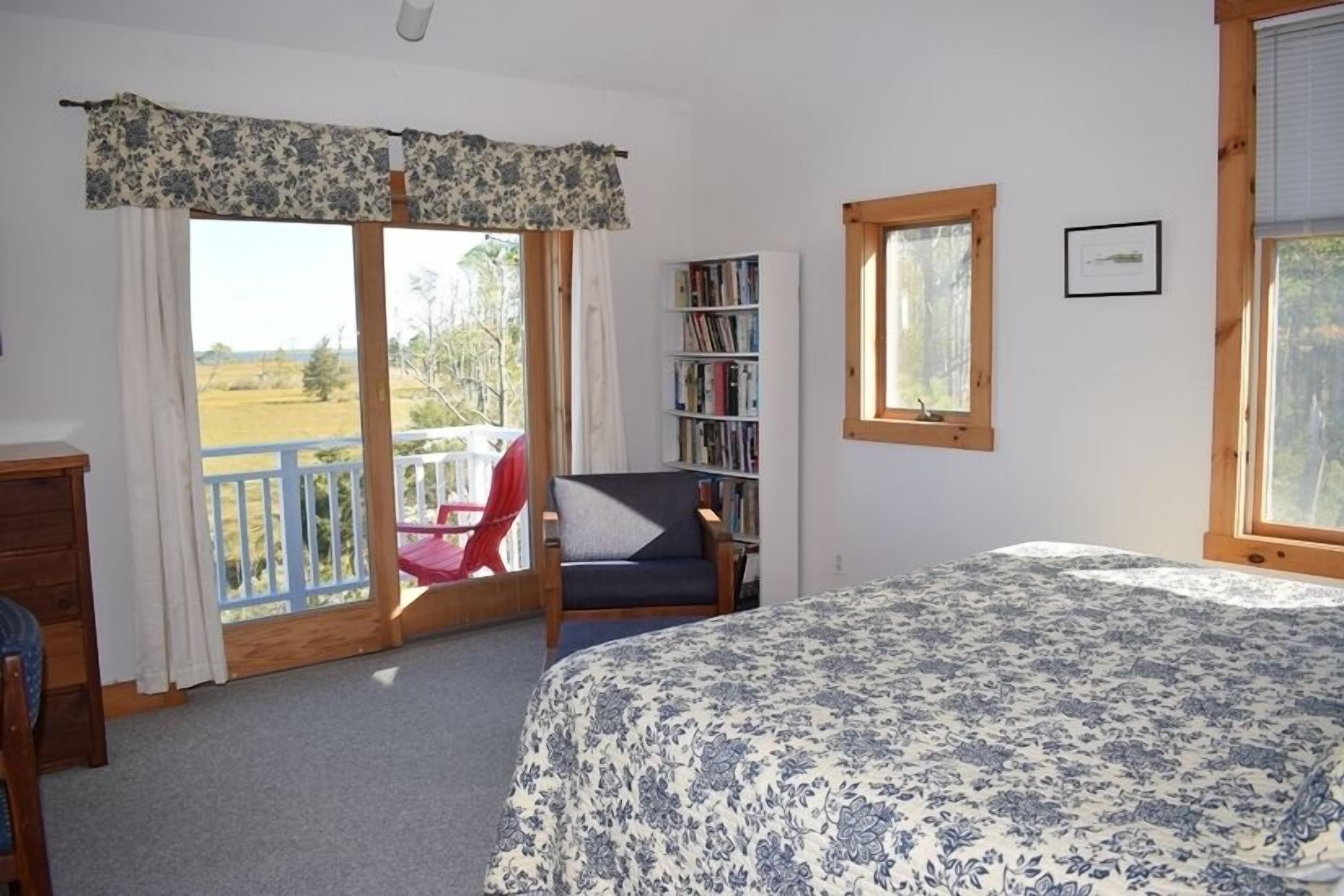 Ein Schlafzimmer mit einem großen Bett, einer Bücherwand, einem Sessel und einem Balkon mit Blick auf die Landschaft.