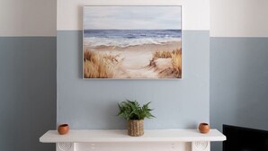 A framed painting of a beach scene on a wall above a white mantelpiece with two small terracotta pots and a woven basket with green plants.