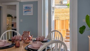 A dining area with a table set for four, white chairs, and a view of a wooden deck and garden through a glass door.