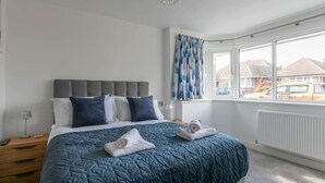 Ein Schlafzimmer mit einem großen Bett, einem grauen Kopfteil und Blick ins Freie durch ein Fenster.