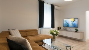 Un salon moderne avec un canapé marron, une table basse en verre, un téléviseur fixé au mur et une console blanche agrémentée d’objets décoratifs.