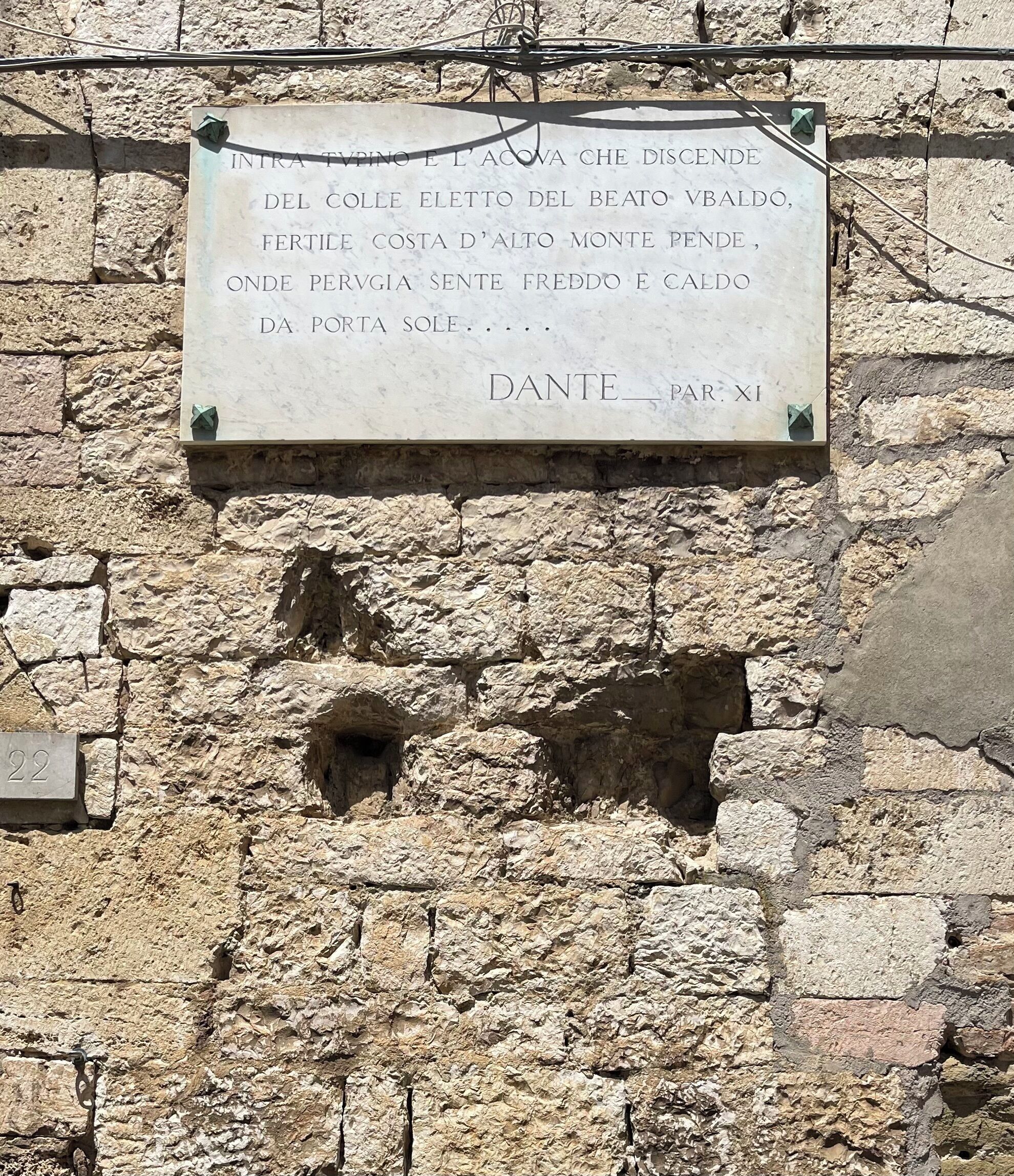 Un mur de pierre sur lequel est fixée une plaque portant l’inscription suivante : « INTRA TAVINO E L’ACOVA CHE DISCENDE DEL COLLE ELETTO DEL BEATO VBALDO, FERTILE COSTA D’ALTO MONTE PENDE, ONDE PERVIA SENTE FREDDO E CALDO DA PORTA SOLE. »
