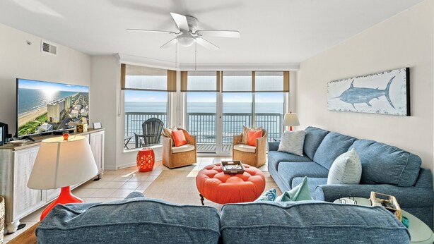 A spacious living room with a large TV, comfortable seating, and a balcony with a view of the ocean.