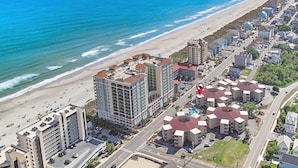 Vista aérea de una zona hotelera costera con varios edificios, incluyendo un rascacielos destacado y varias construcciones más pequeñas con tejados rojos, junto a una playa de arena y el océano.