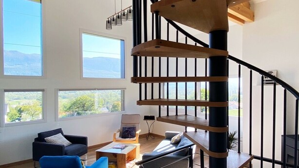 Ein modernes Interieur mit einer Wendeltreppe, Holzbalken und großen Fenstern mit Blick ins Freie.