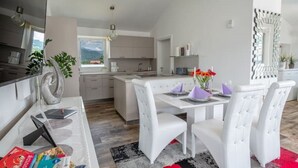Una cocina moderna con armarios blancos, un comedor con sillas blancas y vistas a las montañas a través del ventanal.