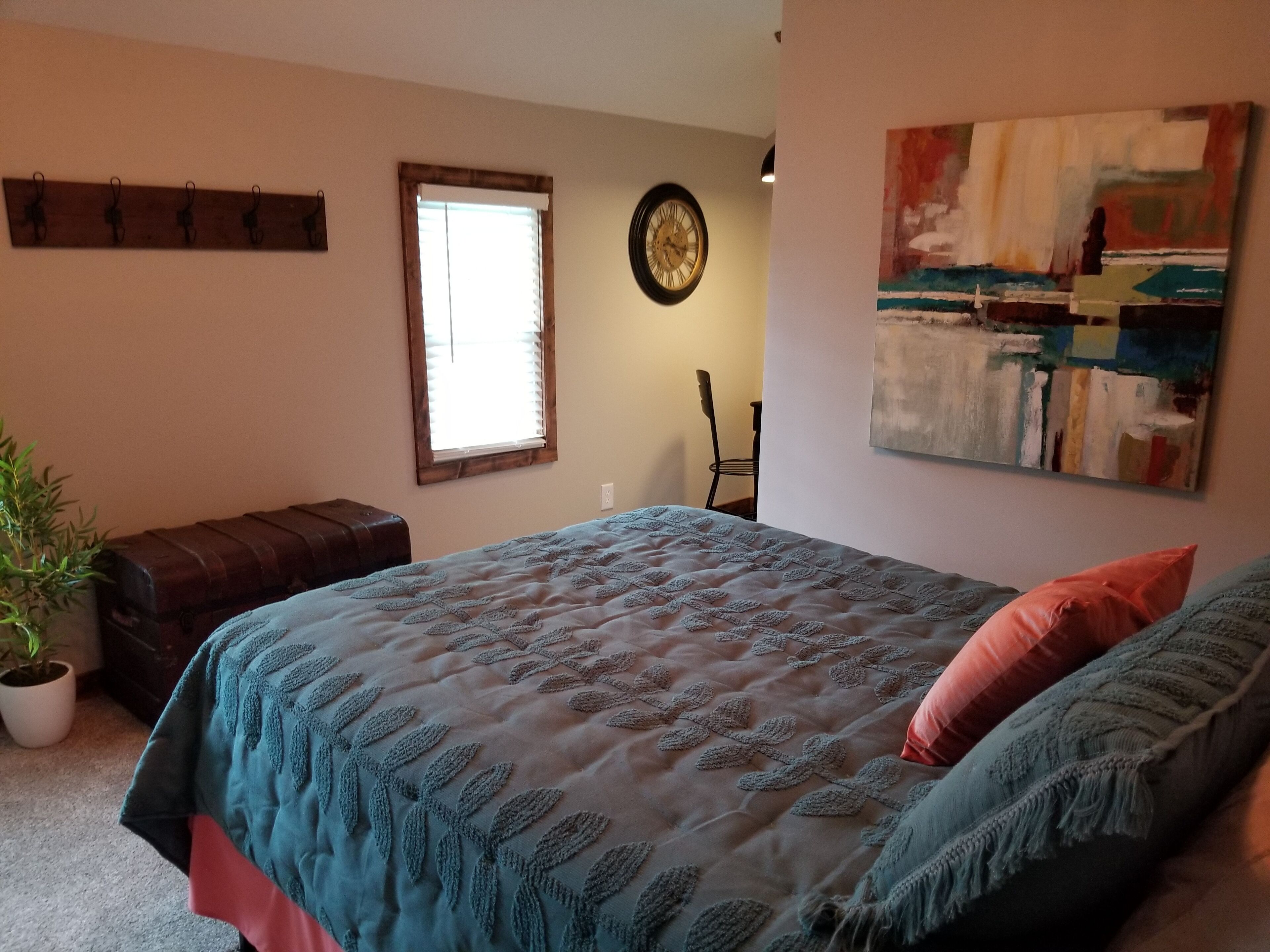 A bedroom with a bed, a bench, a potted plant, a window, a clock, and a painting.