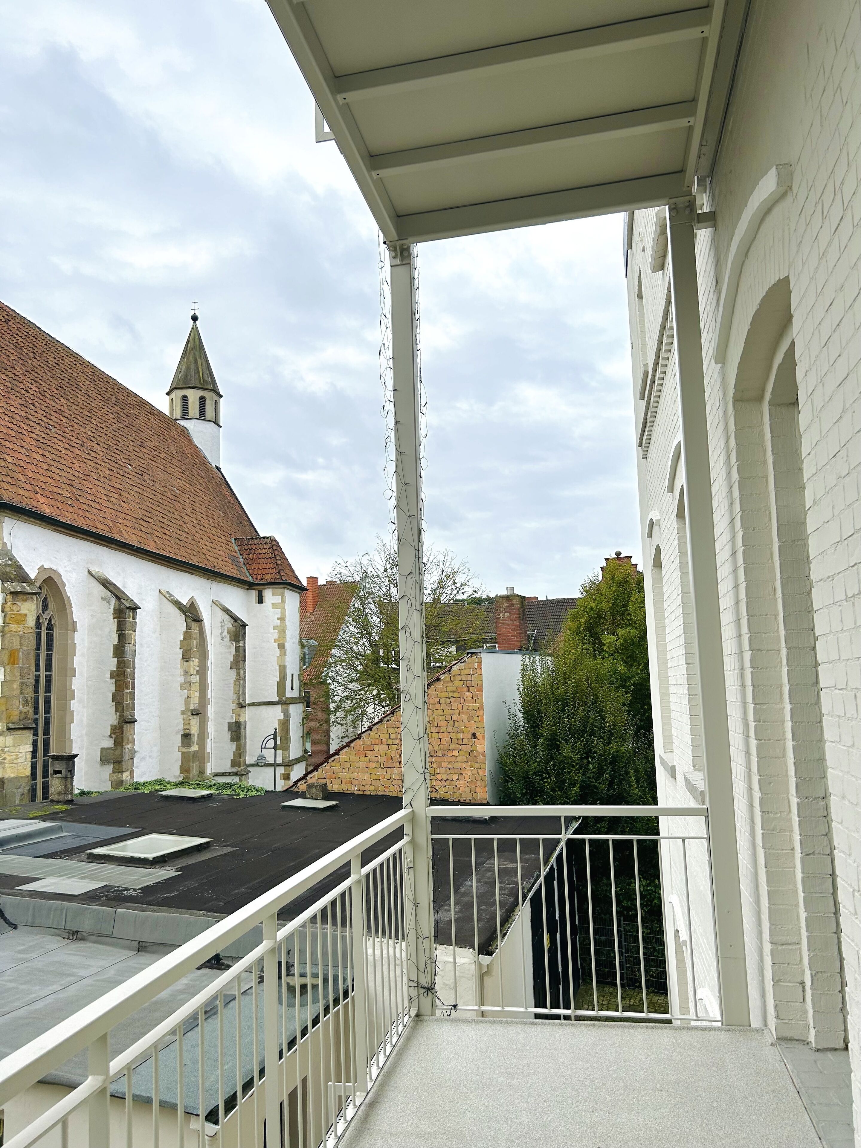 A balcony with a white railing overlooking a church with a red-tiled roof and a tower.