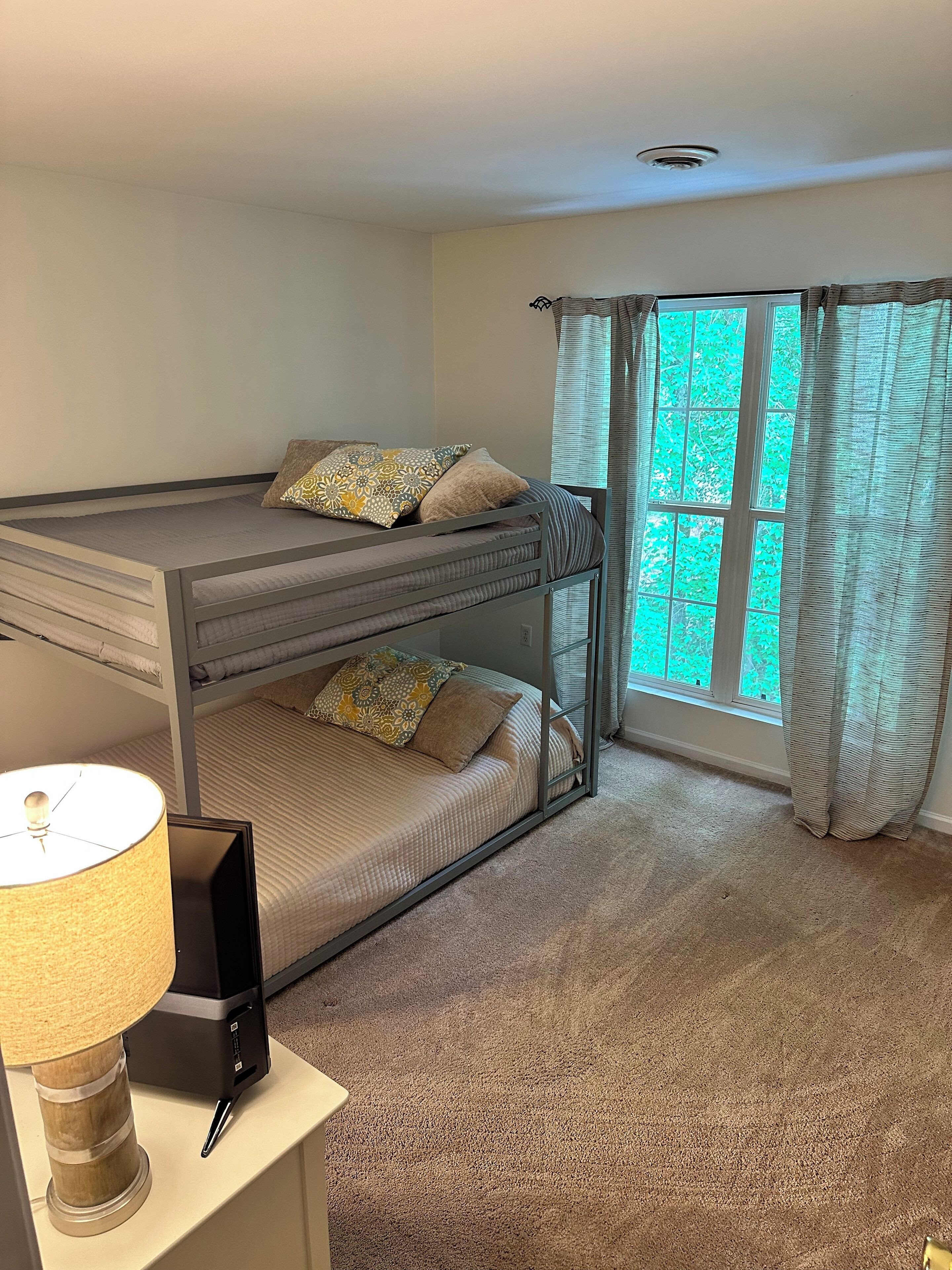 Quarto com beliche, escrivaninha e luminária, amplas janelas com cortinas e piso coberto por carpete.