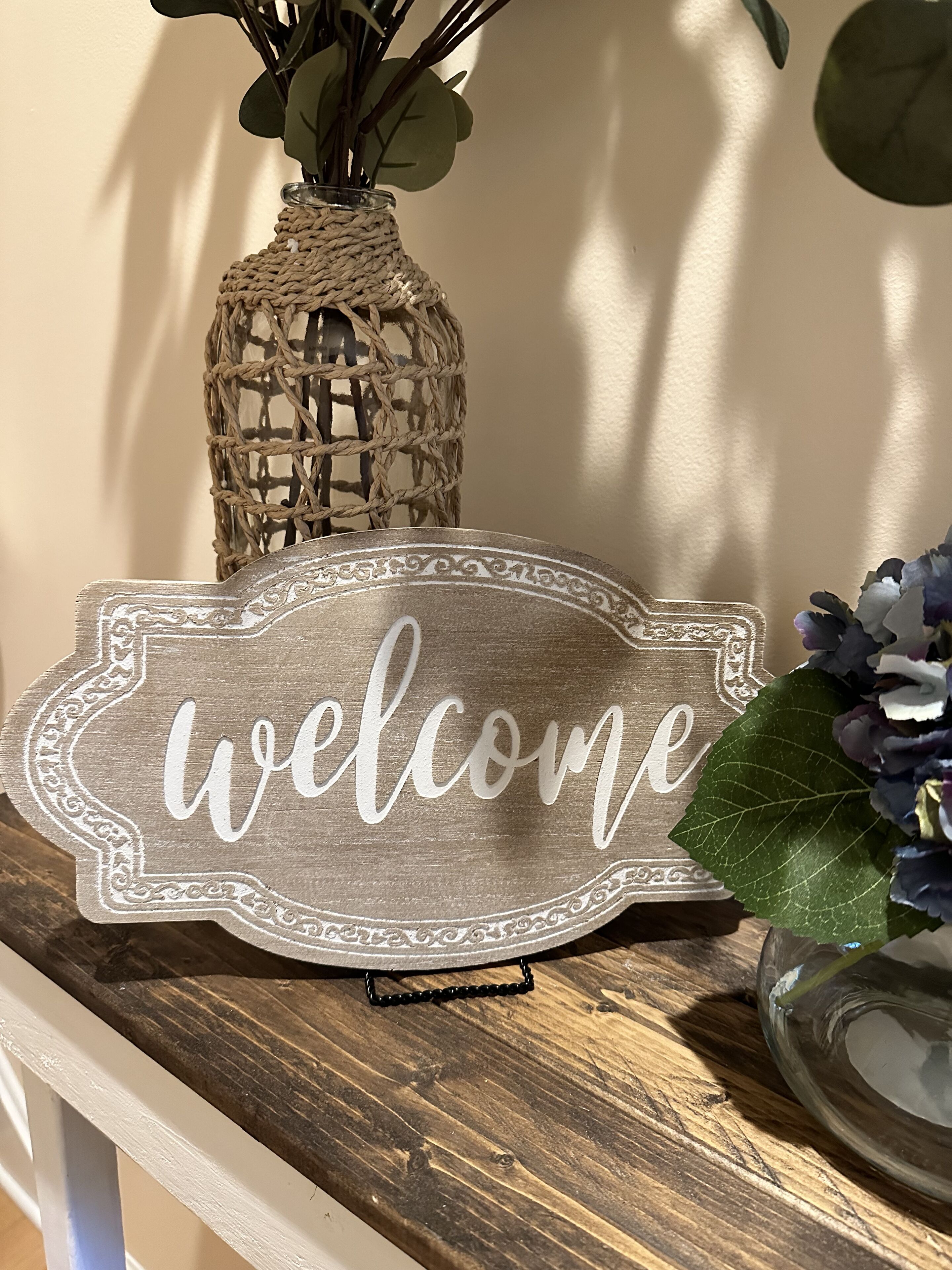 A welcome sign on a wooden surface with a woven basket and flowers in the background.