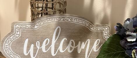 A welcome sign on a wooden surface with a woven basket and flowers in the background.