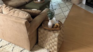 A living room with a beige sofa, a green ottoman, a woven basket, and a potted plant.