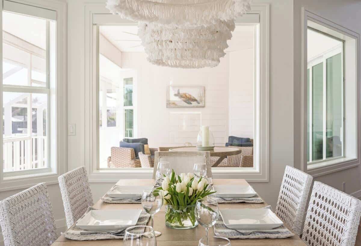 Comedor con una mesa dispuesta para cuatro personas, sillas blancas, un florero y una araña.