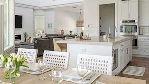 A modern kitchen with white cabinetry, a central island, and a dining area with white wicker chairs. - Gorgeous Waterfront Home; WALK to Beach and shops (Marco Island)
