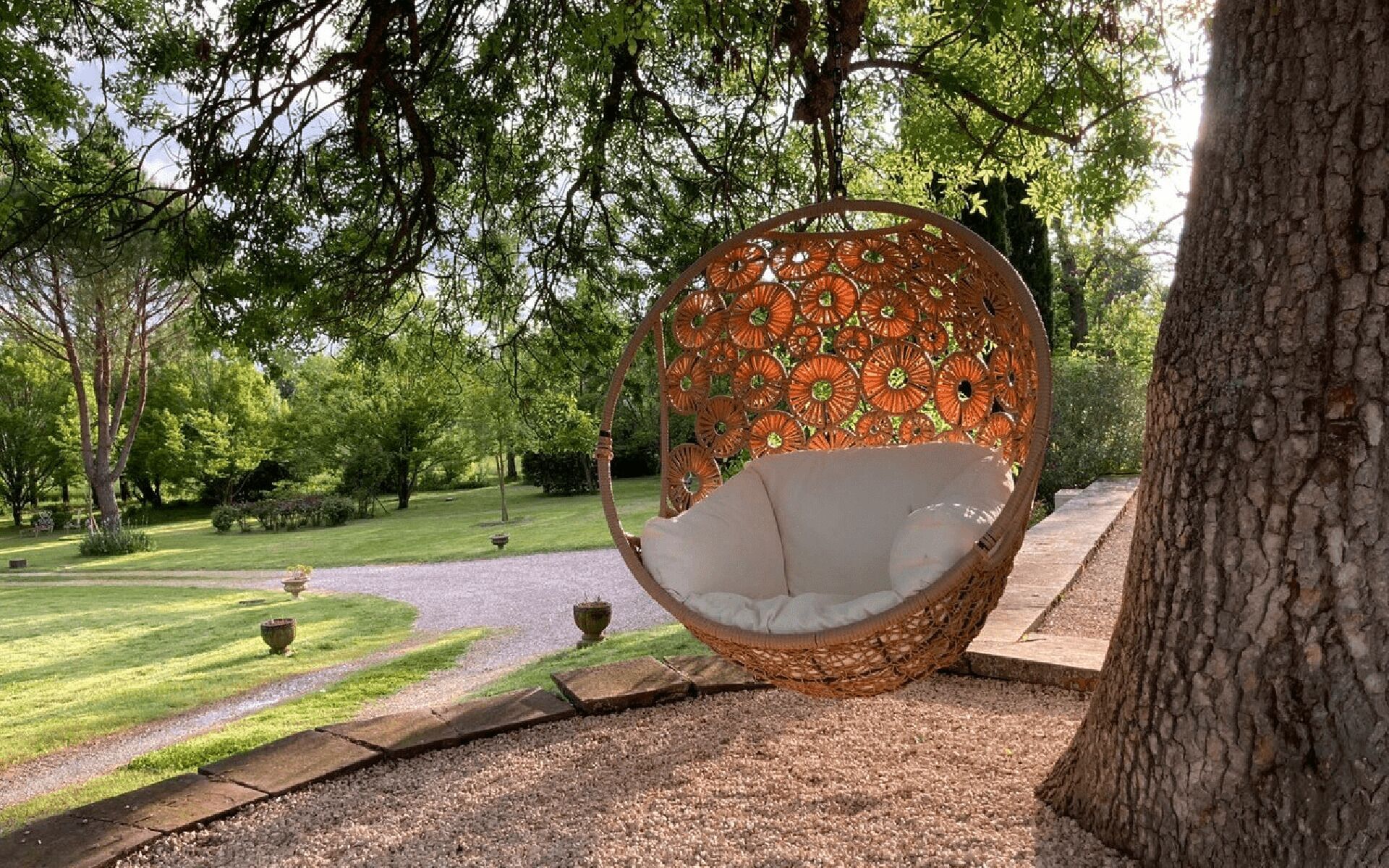 A wicker swing chair with a cushion, positioned under a tree in a park with a gravel path and greenery.