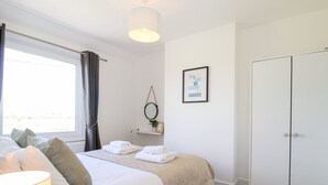 Un dormitorio con cama, lámpara de noche, ventana con cortinas, espejo montado en la pared, una cita enmarcadas, estante con planta y armario.