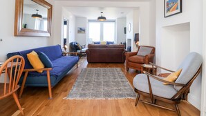 Una sala moderna con un sofá azul, un sofá marrón y un sillón gris. Hay una alfombra estampada en el suelo, una mesa de centro de madera y una silla de madera. Un espejo grande cuelga de la pared, y un cuadro está colgado sobre el sillón.