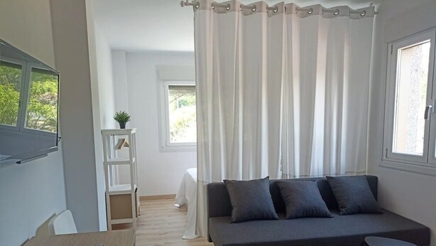 Een moderne woonkamer met een grijze bank, twee zwarte krukken, een houten tafel en een witte plank met een plant.