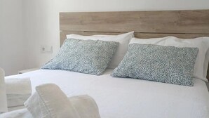 Een netjes opgemaakt bed met witte lakens en gekleurde kussens, een houten hoofdbord en een stapel witte handdoeken op de vloer.