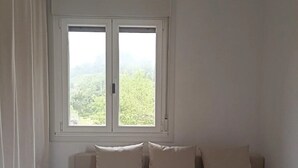 A minimalist room with a sofa, two green chairs, a window with curtains, and a view of greenery outside. - Casal do Crego (O Grove)
