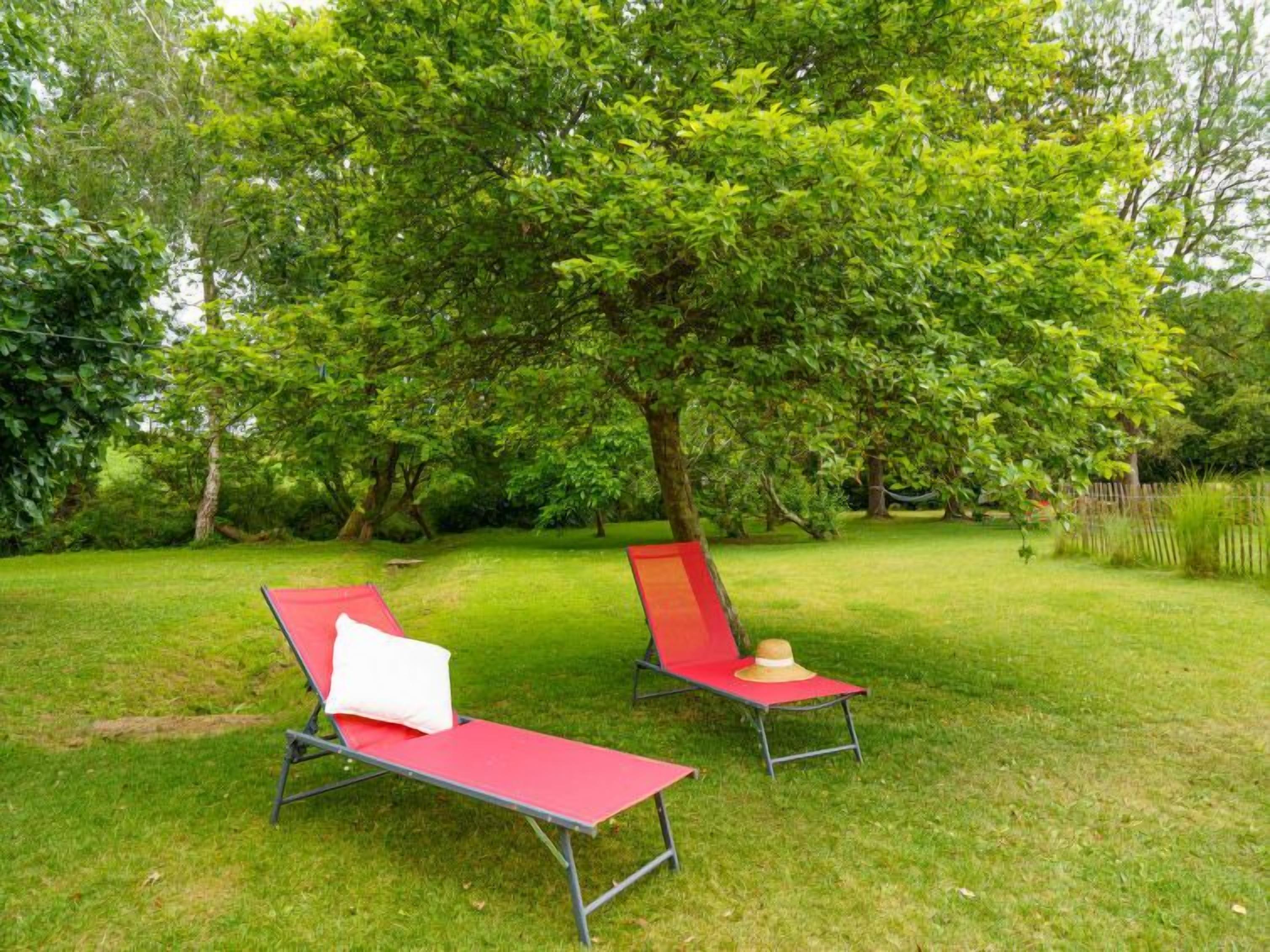 Two red lounge chairs with white cushions and a straw hat on a grassy lawn under a large tree.