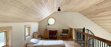A loft bedroom with a sloped ceiling, wooden beams, a desk, two beds, a chair, and a window.