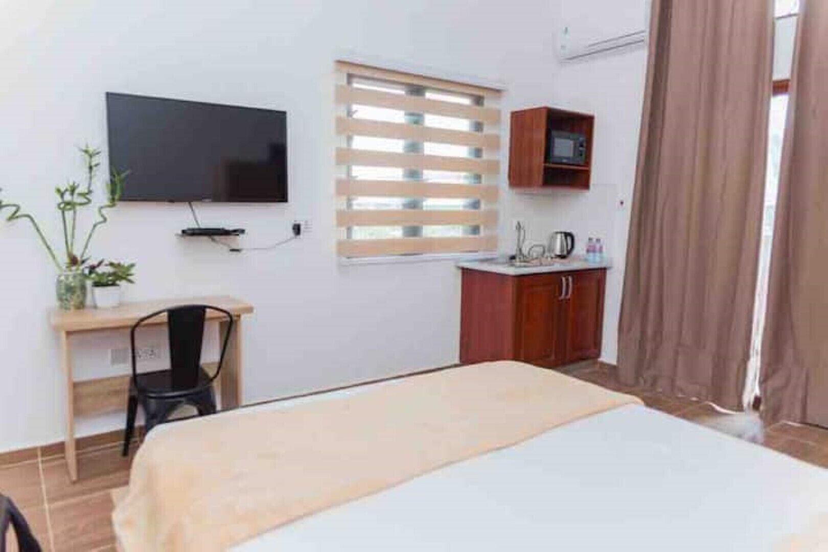 Ein Schlafzimmer mit einem Bett, einem Schreibtisch mit Stuhl, einem Fernseher, einem Fenster mit Jalousien, einer Mikrowelle und einem Schrank.