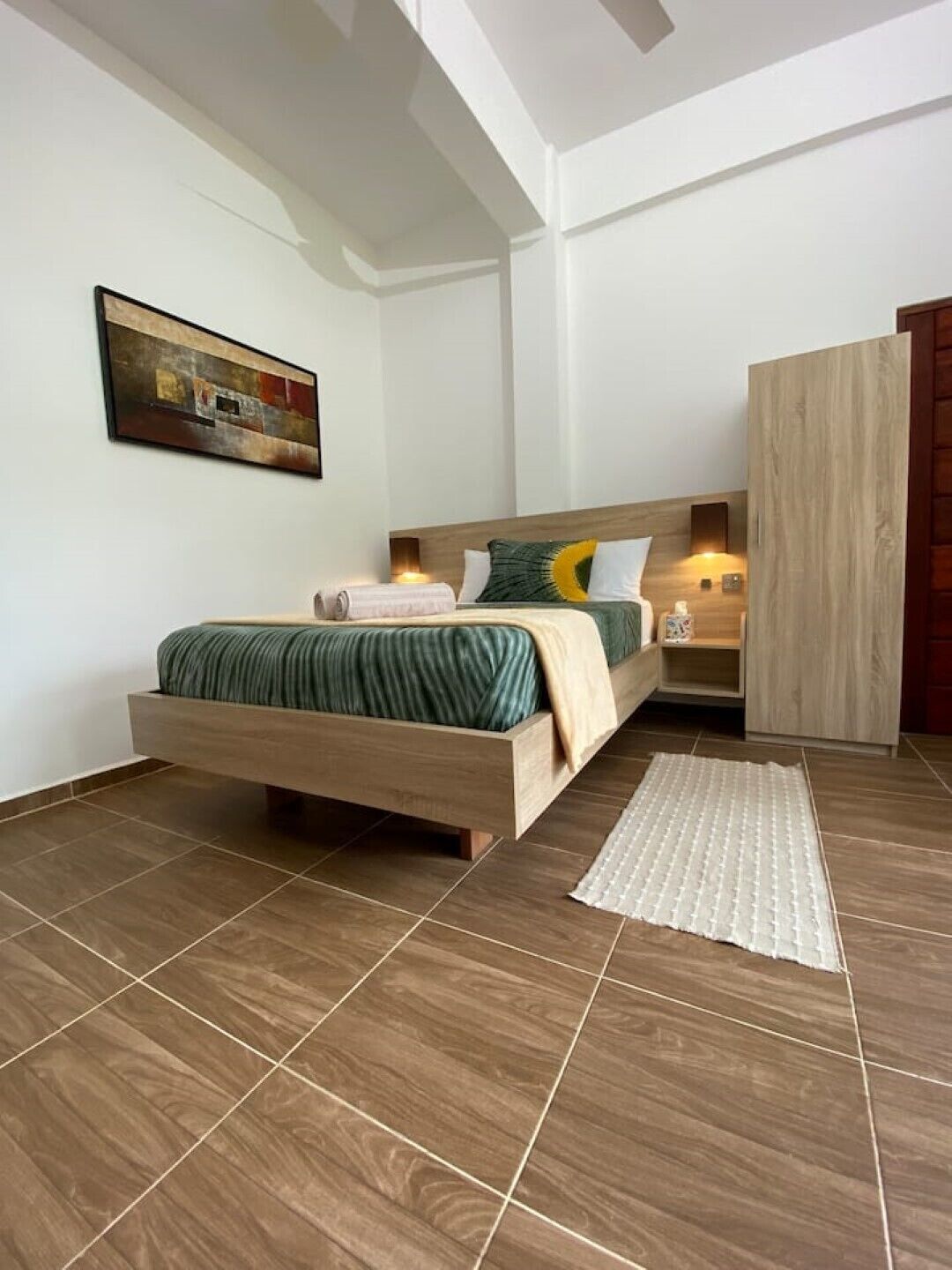 A modern bedroom with a wooden bed, a nightstand, a wall-mounted lamp, and a painting on the wall.