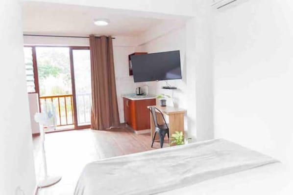 A hotel room with a bed, a desk, a chair, a TV mounted on the wall, and a sliding glass door leading to a balcony.