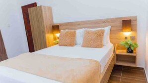 A neatly made bed with a wooden headboard, two bedside tables with lamps, and a small plant on one table. - The Treehouse Studio apartments with a large rooftop terrace. (Aburi-South)
