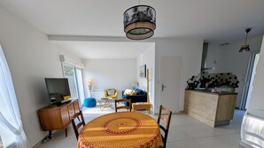 A dining area with a round table, chairs, and a patterned tablecloth. There is a television on a cabinet to the left, and a kitchen area with a wooden countertop and a hanging light fixture.
