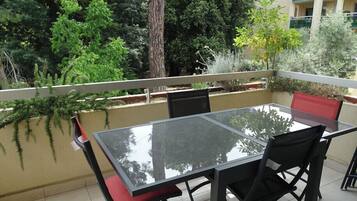 A balcony with a glass table and chairs, surrounded by greenery and a building in the background.