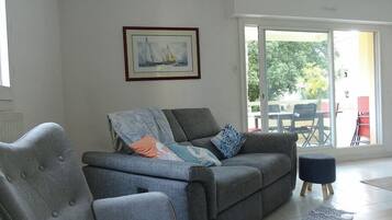 Un salon avec un canapé gris, un fauteuil, un tableau encadré au mur et une vue sur une terrasse extérieure avec des chaises et une table.