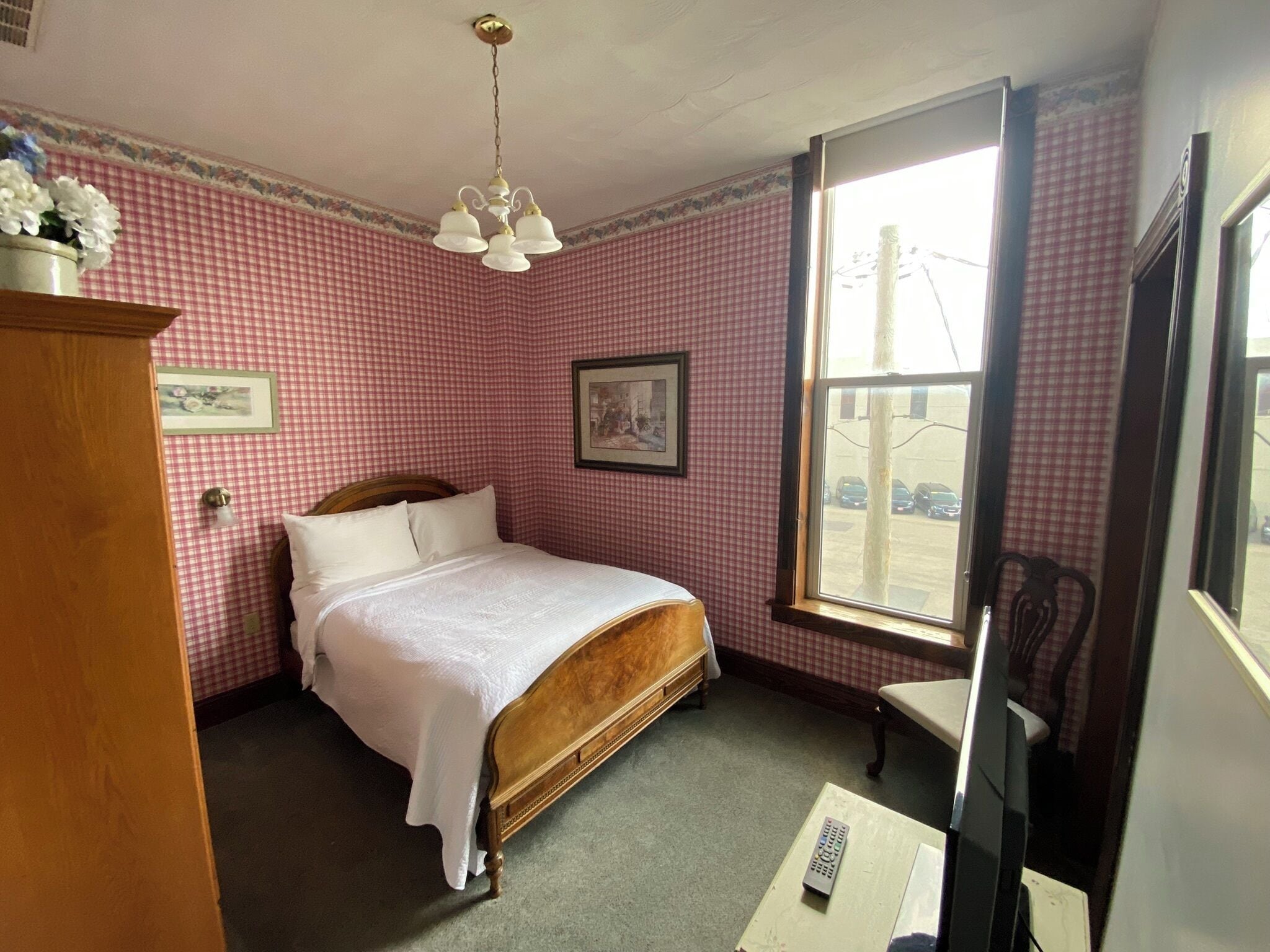 A bedroom with a wooden bed, a window with a view of parked cars, a chair, and a dresser.