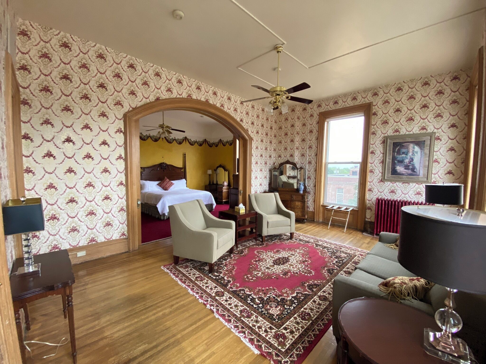 Habitación de hotel con cama, dos sillones, una mesa de centro, alfombra roja estampada, ventilador de techo y un ventanal con vista a la ciudad.