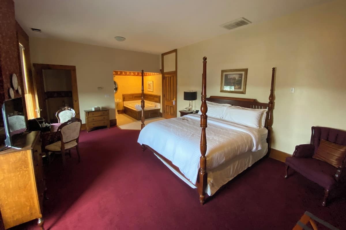 A four-poster bed with a wooden frame, a sitting area with a chair and a small table, a desk with a chair, a large mirror, and a painting on the wall.