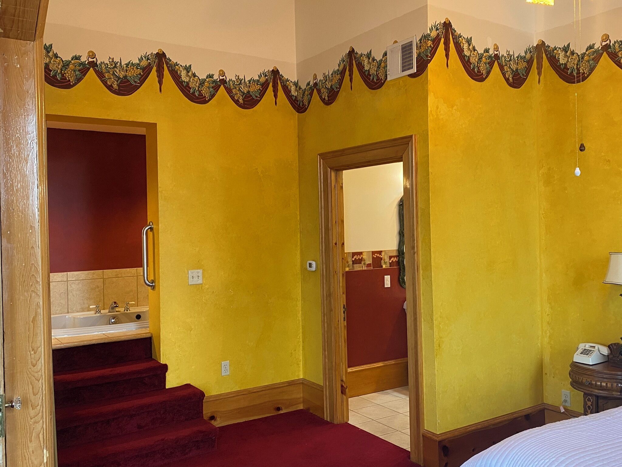 A hotel room with yellow walls, a red carpeted staircase, a wooden door, and a bed with a white comforter.
