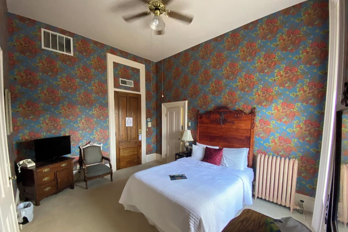 A bedroom with floral wallpaper, a wooden headboard, a bed with white linens, a nightstand, a chair, a desk with a telephone, and a ceiling fan.