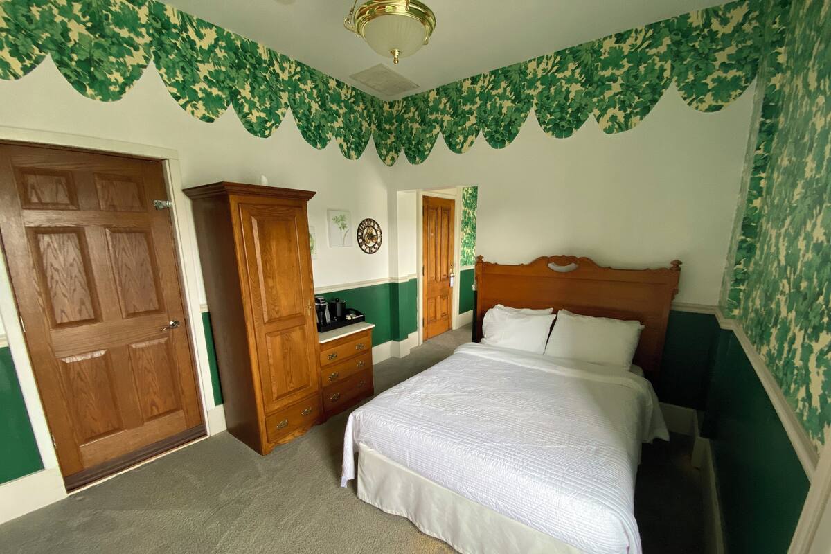 A bedroom with a wooden wardrobe, a bed with white bedding, and green patterned wallpaper.