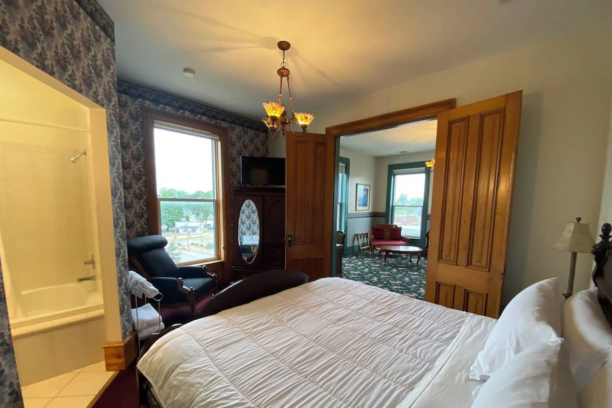 A hotel room with a large bed, a sitting area with a chair and a table, a bathroom with a shower, and a view of the outside through a window.