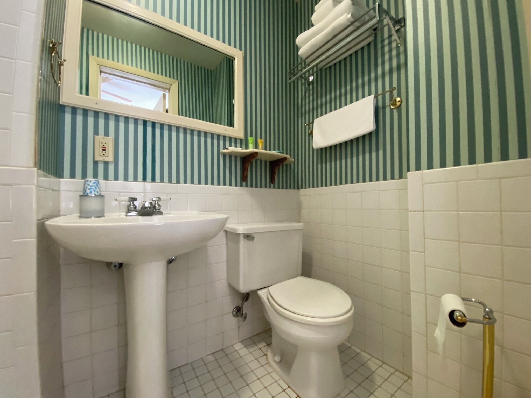 A bathroom with a white sink, toilet, and a mirror. There is a towel rack above the toilet and a shelf with toiletries.