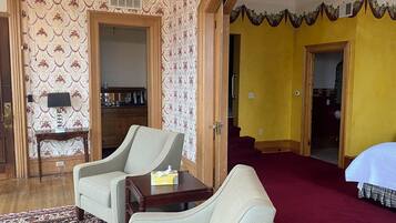 A hotel room with a wooden floor, two armchairs, a small table, a patterned wallpaper, a yellow accent wall, and a red carpet.